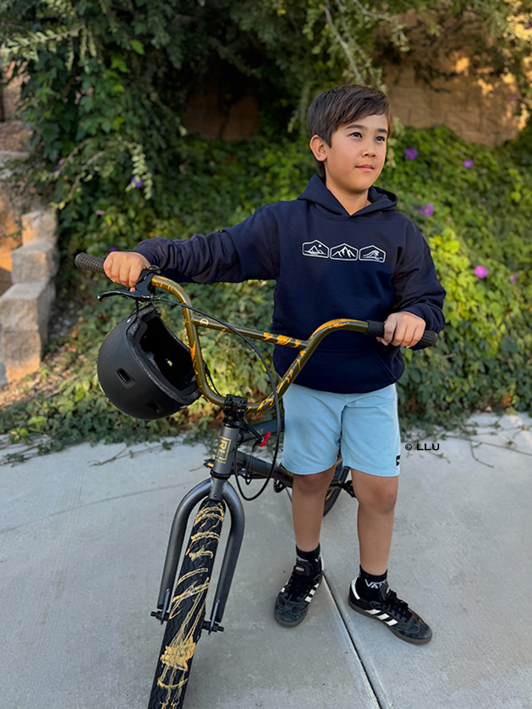 Child holding a bicycle with a helmet, standing outdoors, wearing desert moutain, and ocean graphics