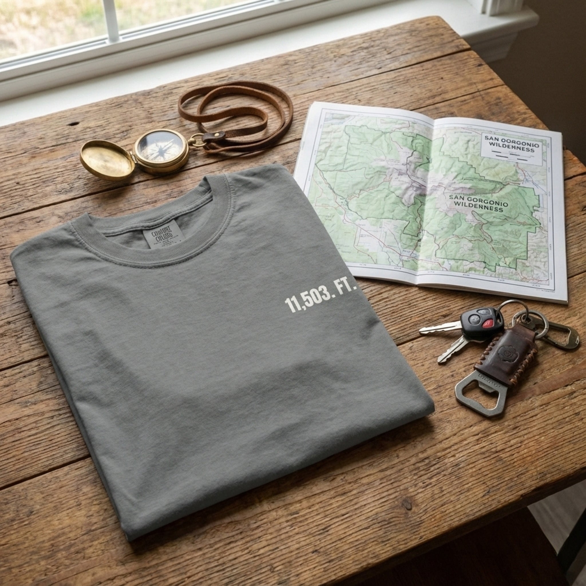 Folded gray t-shirt with '11,503 FT.' on a wooden table with map, keys, and compass.