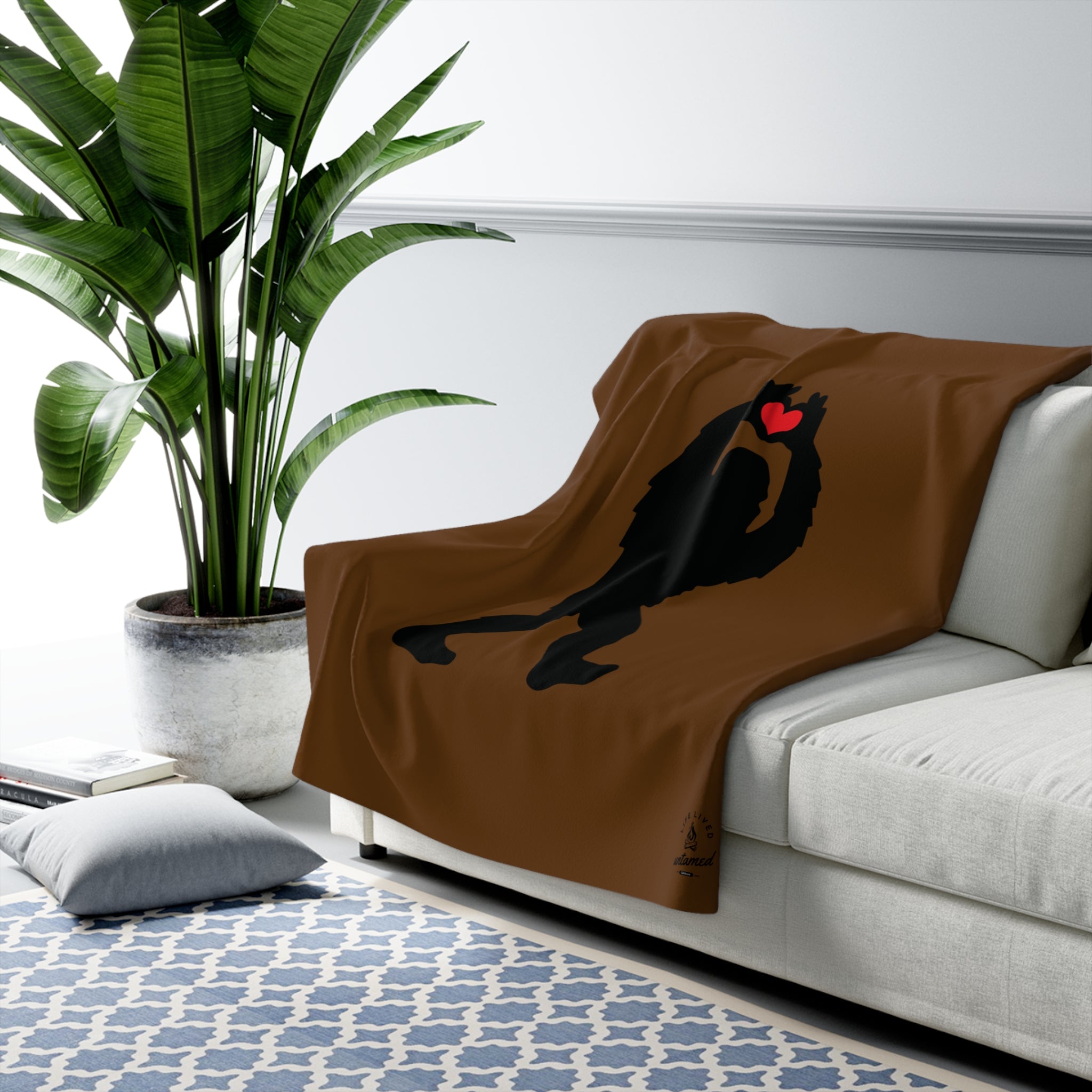 A brown blanket with a black silhouette of a Sasquatch and red heart on a white couch, next to a potted plant.