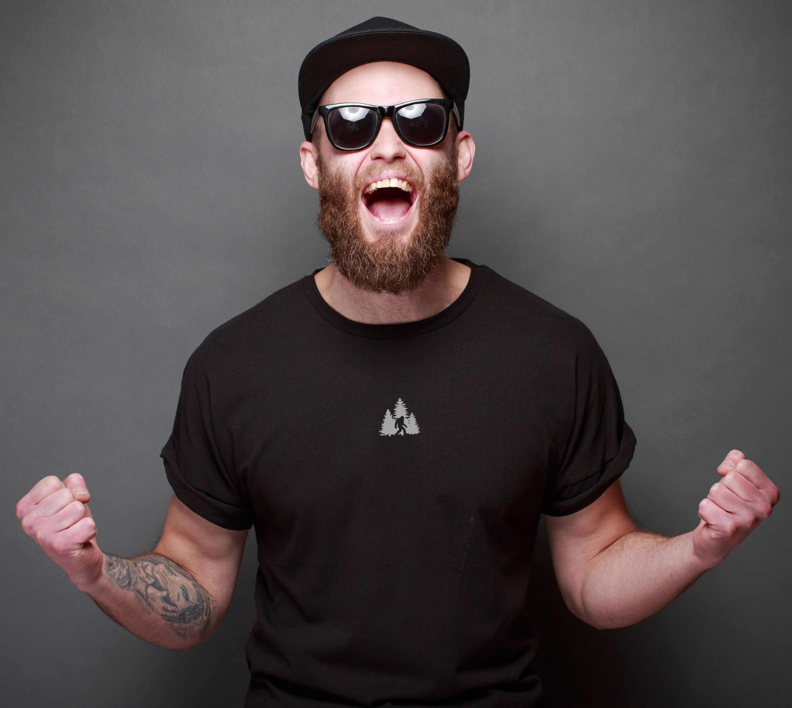 A man wearing sunglasses, black cap, and black t-shirt with a Sasquatch in wilderness graphic on.