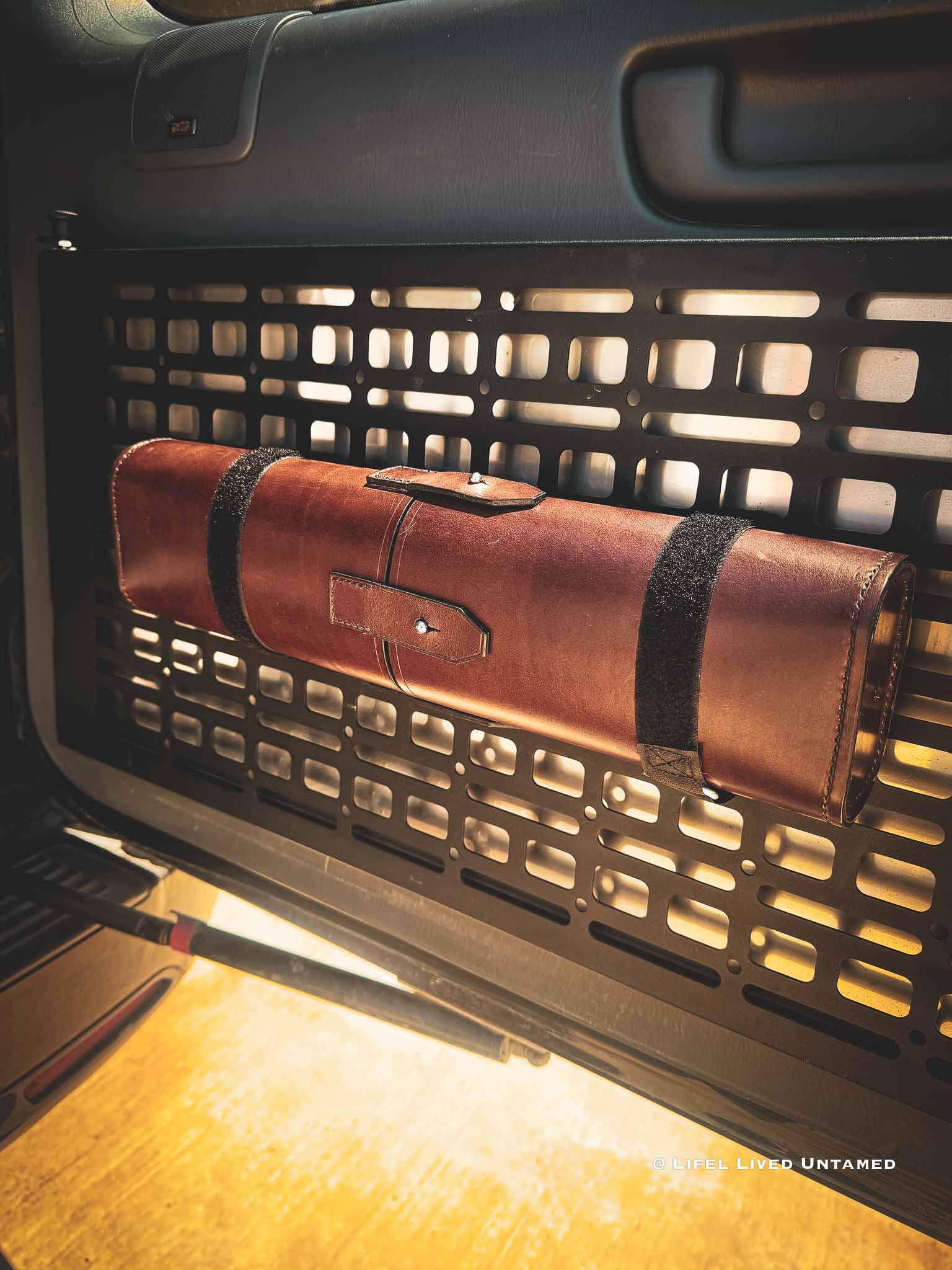 Brown leather tool-roll mounted on molle panel with black Velcro straps inside a vehicle's open door