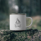 White enamel mug with campfire design on a log in a forest setting.