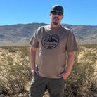 A man standing in a sunny, arid desert landscape with mountains in the background. He is wearing espresso color t-shirt with a black circular logo on the chest that features mountains and the text 'LIFE LIVED UNTAMED', along with dark green cargo pants, sunglasses, and a dark baseball cap. He has his hands in his pockets.