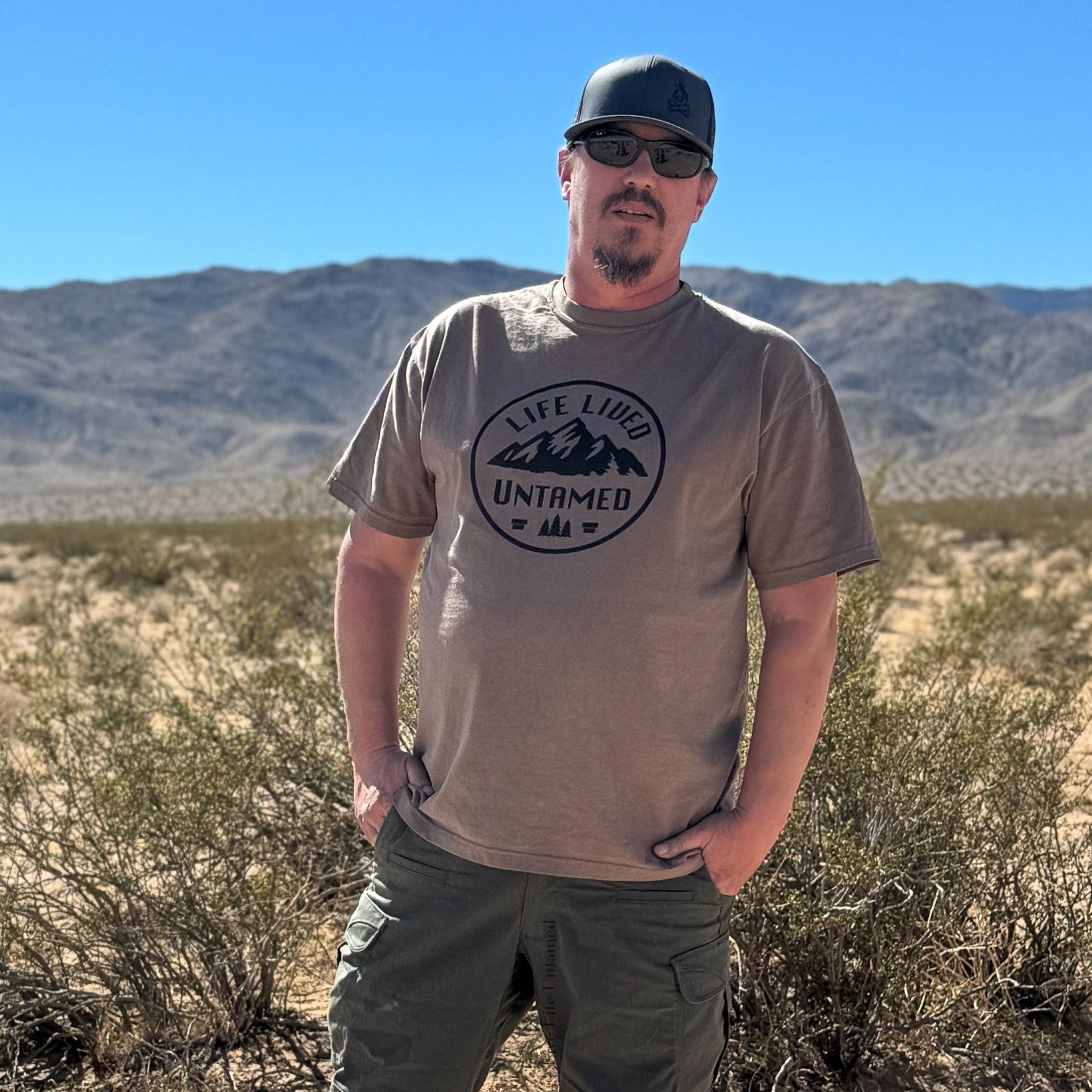 A man standing in a sunny, arid desert landscape with mountains in the background. He is wearing espresso color t-shirt with a black circular logo on the chest that features mountains and the text 'LIFE LIVED UNTAMED', along with dark green cargo pants, sunglasses, and a dark baseball cap. He has his hands in his pockets.