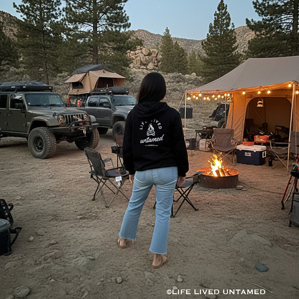 Young standing in the center of a campsite with tents, vehicles, and a fire pit in a forest setting.