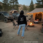 Young standing in the center of a campsite with tents, vehicles, and a fire pit in a forest setting.