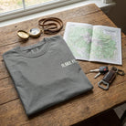 Folded gray t-shirt with '11,503 FT.' on a wooden table with map, keys, and compass.