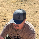 A close-up, high-angle photo of a man wearing a charcoal gray and black mesh trucker hat with a dark embroidered campfire icon on the front. 