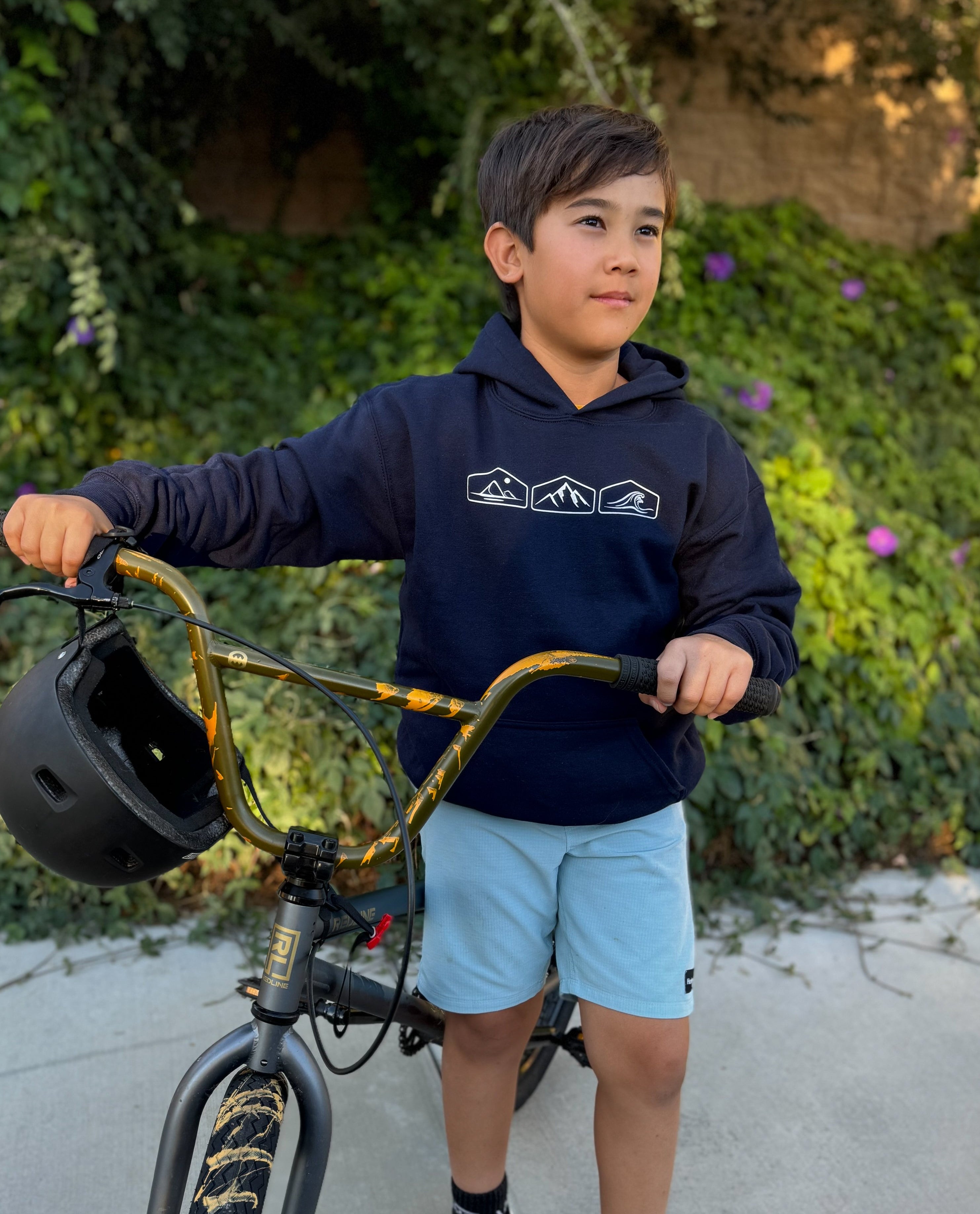A young boy wearing a navy blue hooded sweatshirt with a horizontal row of  mountain, desert, and wave graphics on the front. 